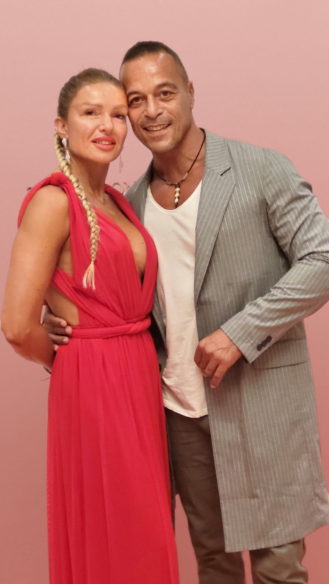 Woman in a red dress and man in a gray suit posing against a pink background.