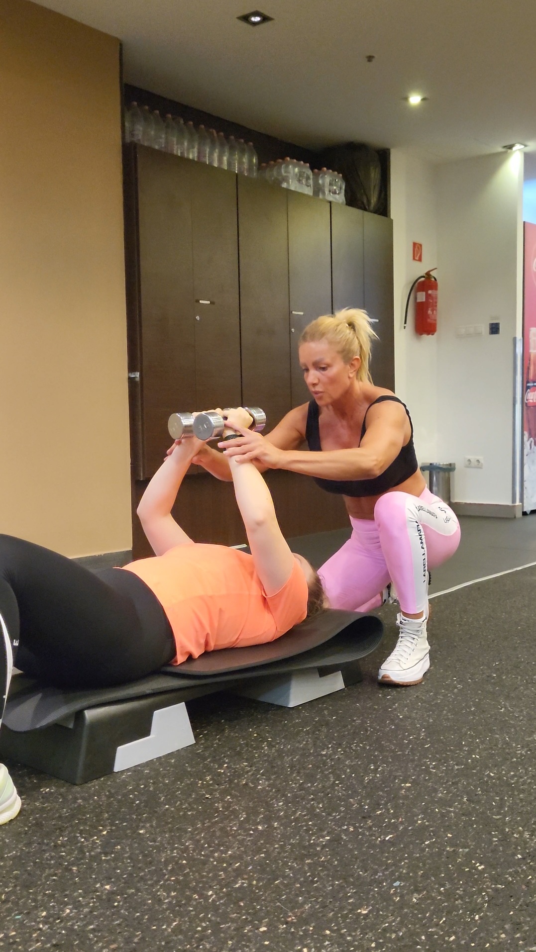 Trainer assisting a person with dumbbell exercise on a gym mat.