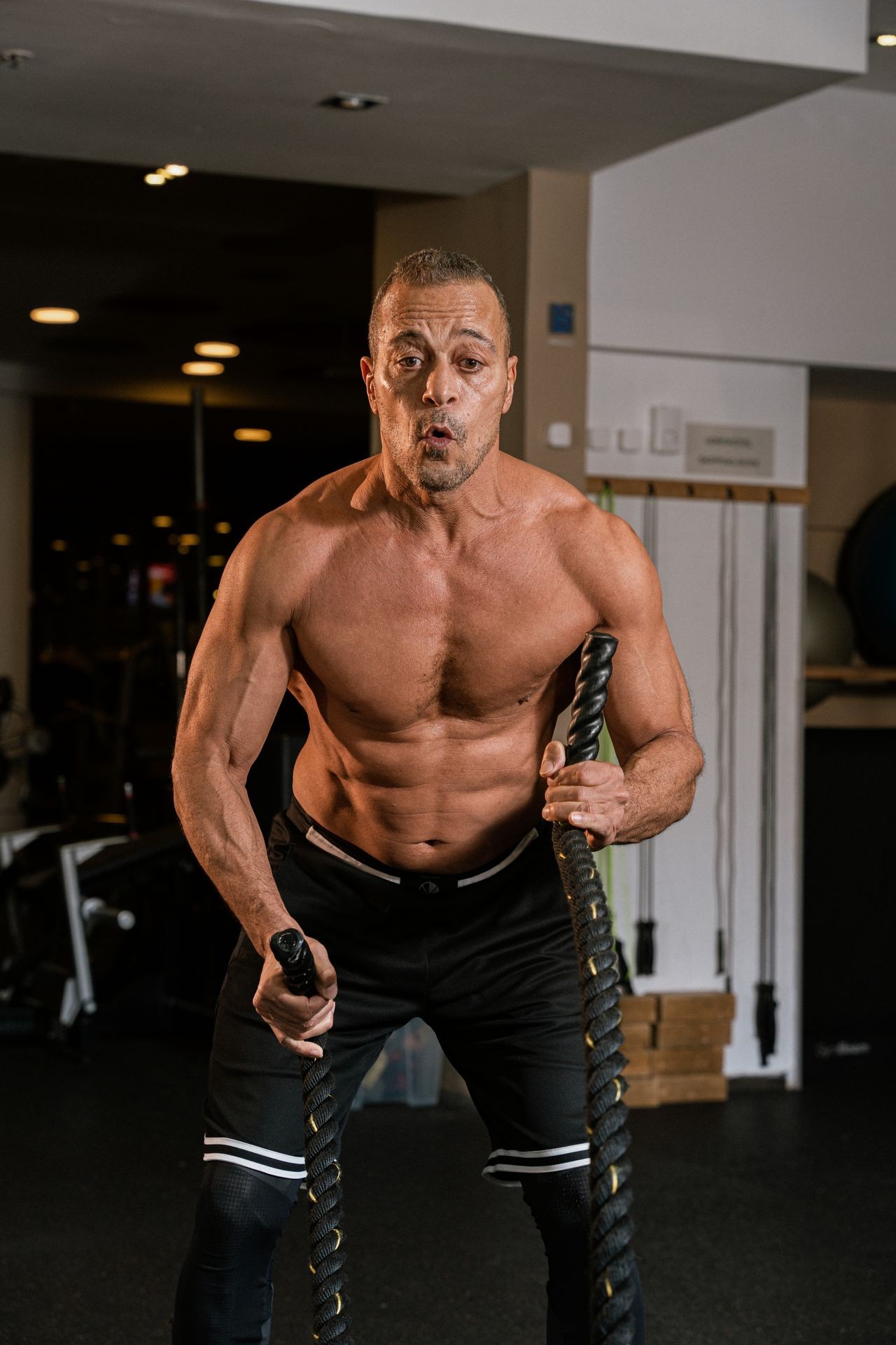 Man working out with battle ropes in a gym, showcasing muscular build and intense focus.